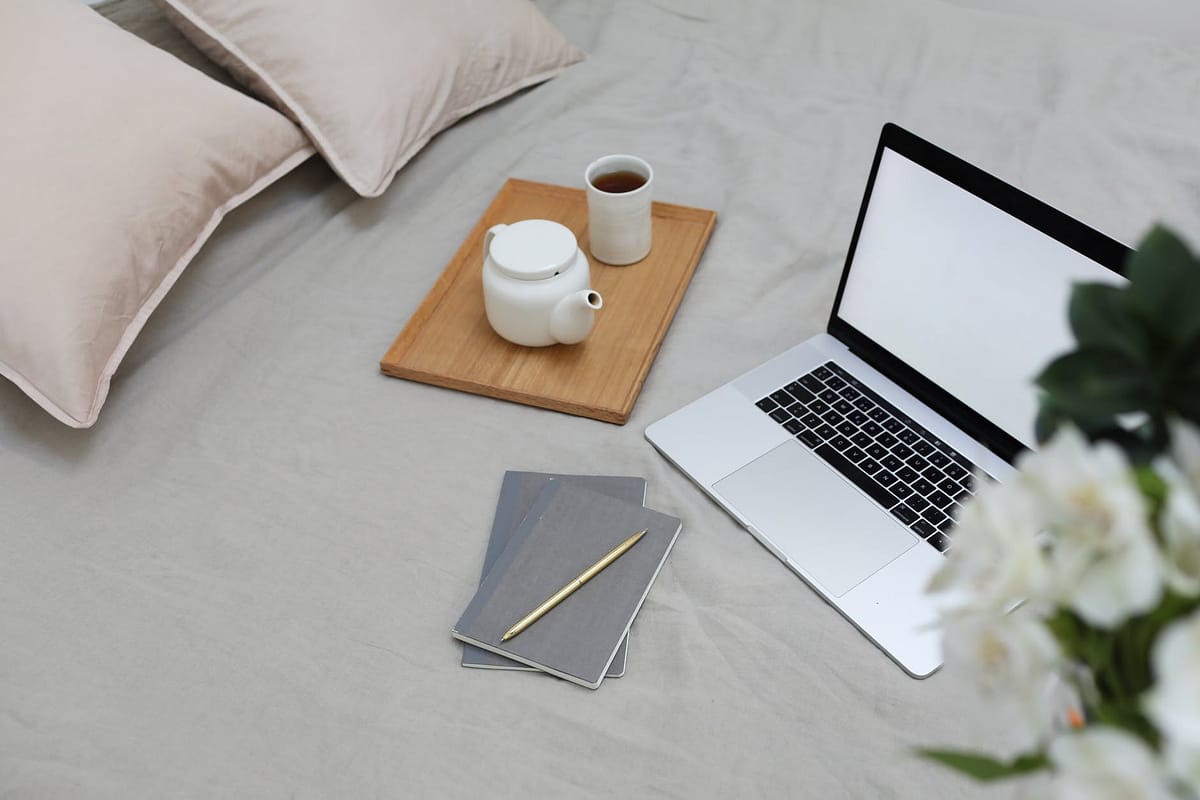 modern laptop with notebooks and pen arranged on bed with coffee pot and cup